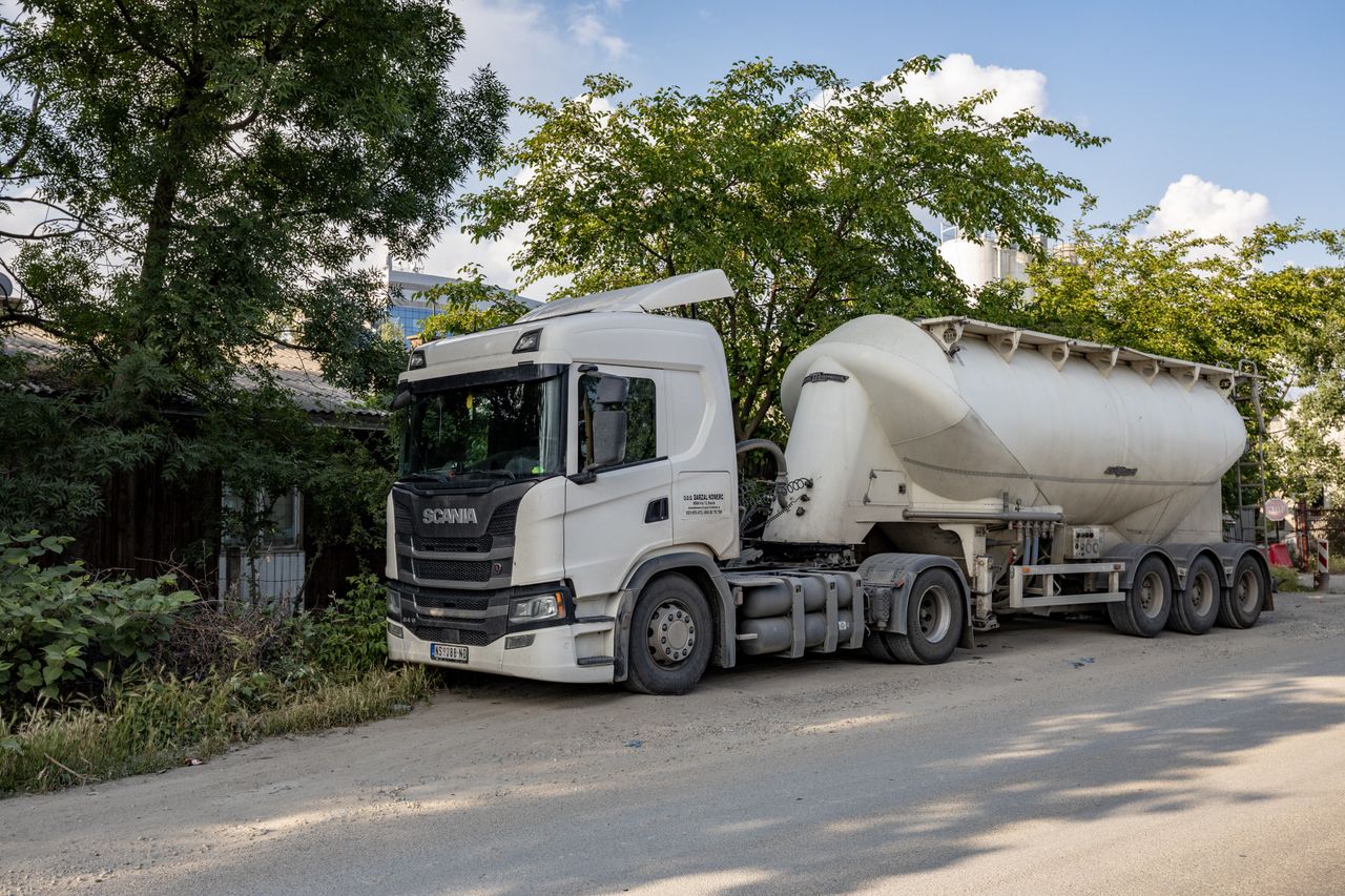 White Scania tanker truck parked by trees on a sunny city street.